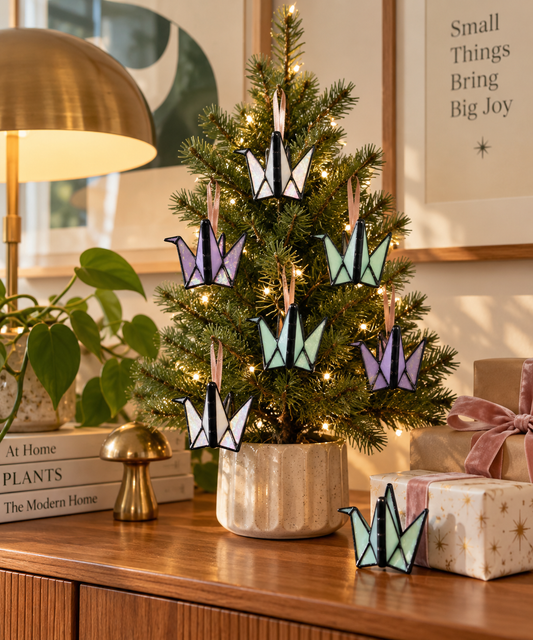 A decorated Christmas tree in a real home setting, with warm lights, wrapped gifts, books, a lamp, and framed wall art. The tree is adorned with multiple origami crane ornaments in pastel purple, mint green, and white, with one matching ornament placed in front on a wooden surface.