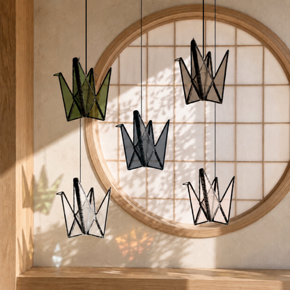 Five handmade stained glass origami crane suncatchers in olive green, smoky blue-gray, taupe, blush, and clear glass hanging in front of a round Japanese-style window in a warm, sunlit Japandi interior.