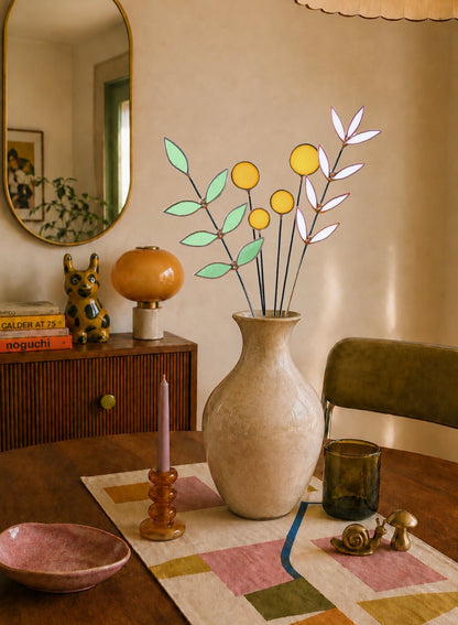 A beige ceramic vase holding a decorative stained glass botanical arrangement with yellow and white flowers and green leaves, placed on a dining table in a warm, styled room with a mirror, side cabinet, and other home decor.