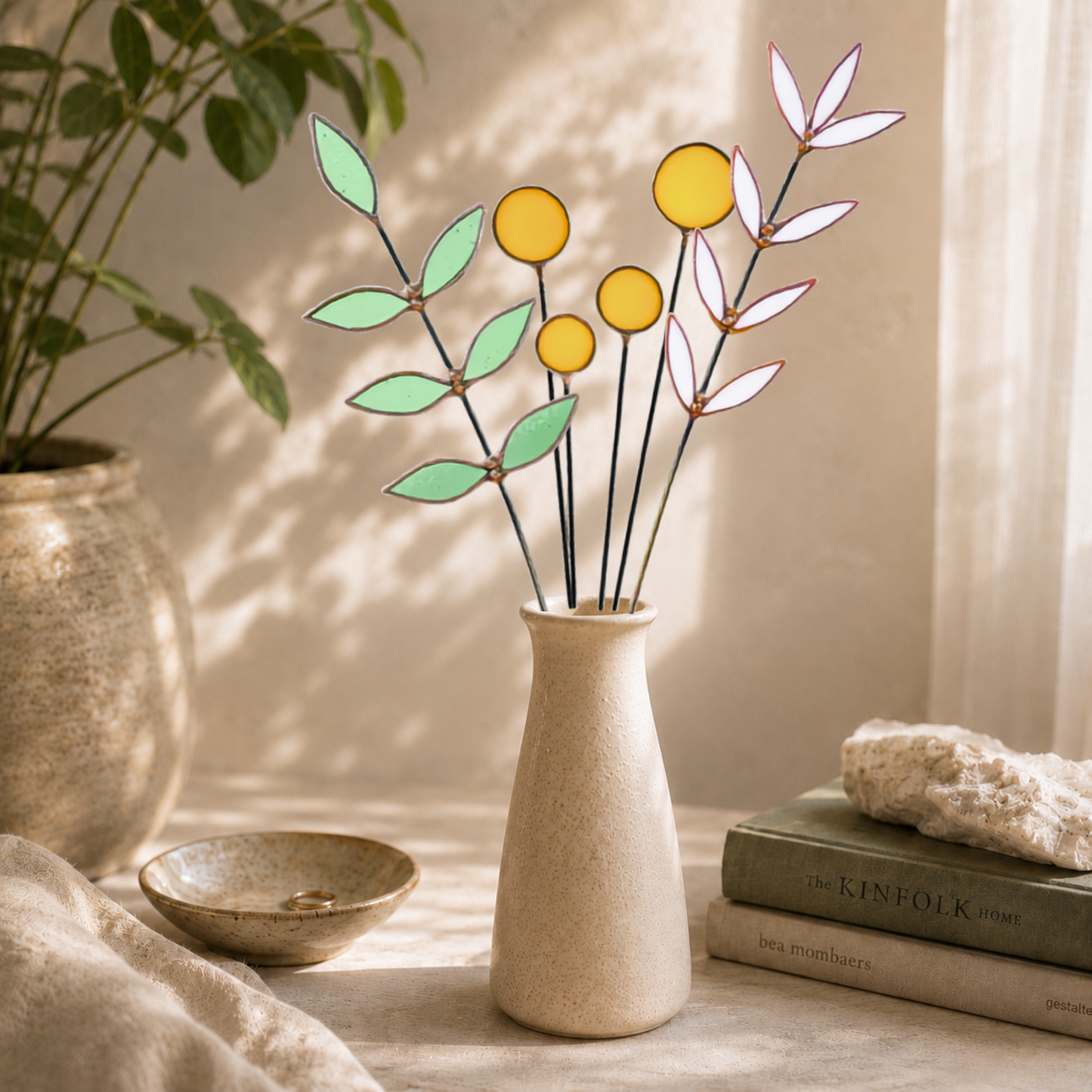 A set of stained glass flower stems with yellow circles and green and white leaves, arranged in a textured beige ceramic vase on a table with a bowl, books, and a plant in the background.