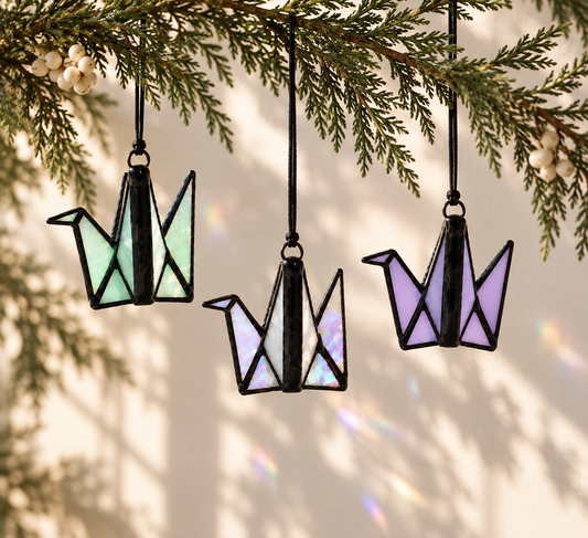Three origami crane ornaments hanging from a pine branch, each made of iridescent stained glass in mint green, white, and lavender, with black metal framing.