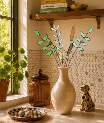 A decorative floral arrangement with green leaves and cattails displayed in a cream ceramic vase on a kitchen counter, with a potted plant, amber glass jar, garlic, and a dog statue nearby.