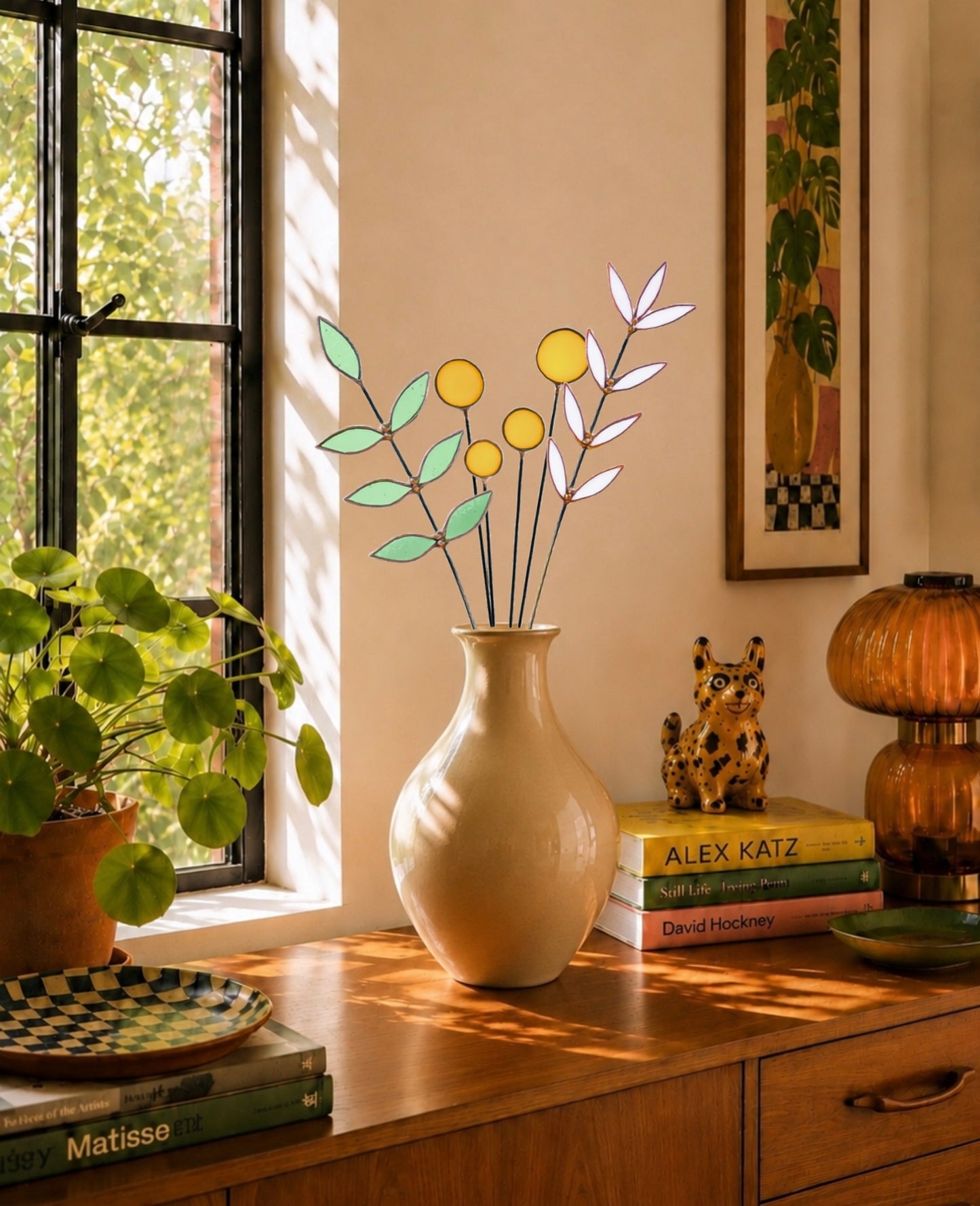 A beige ceramic vase holding a decorative stained glass floral arrangement with pale green leaves and yellow circular blooms, placed on a wooden cabinet in a sunlit room beside a window, books, a plant, and other decor.