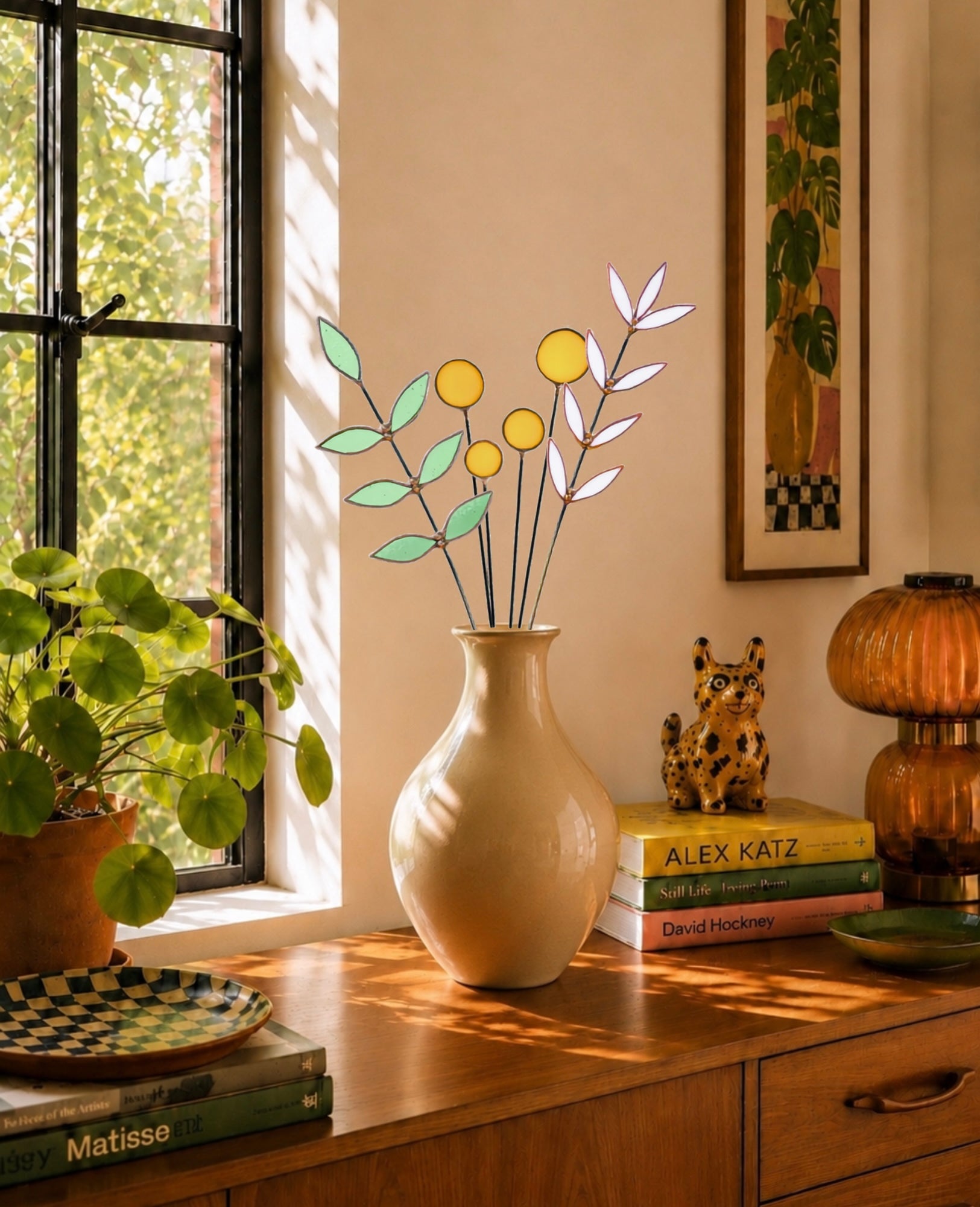 A beige ceramic vase holding a decorative stained glass floral arrangement with pale green leaves and yellow circular blooms, placed on a wooden cabinet in a sunlit room beside a window, books, a plant, and other decor.