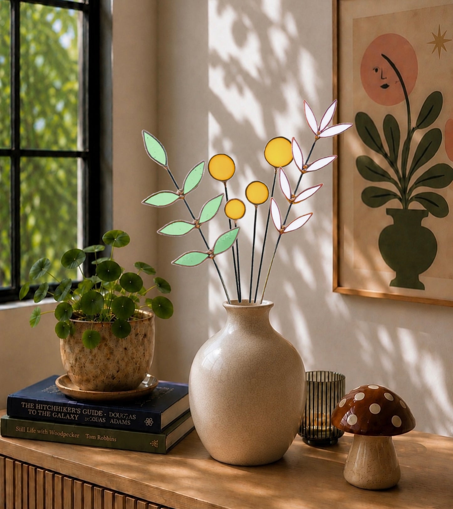 A decorative glass vase arrangement with pale green leaves and yellow circular accents on black stems, displayed in a beige ceramic vase on a wooden table in a sunlit room. A potted plant, books, a small candle holder, and a mushroom-shaped decor piece are nearby, with a framed botanical print on the wall behind.
