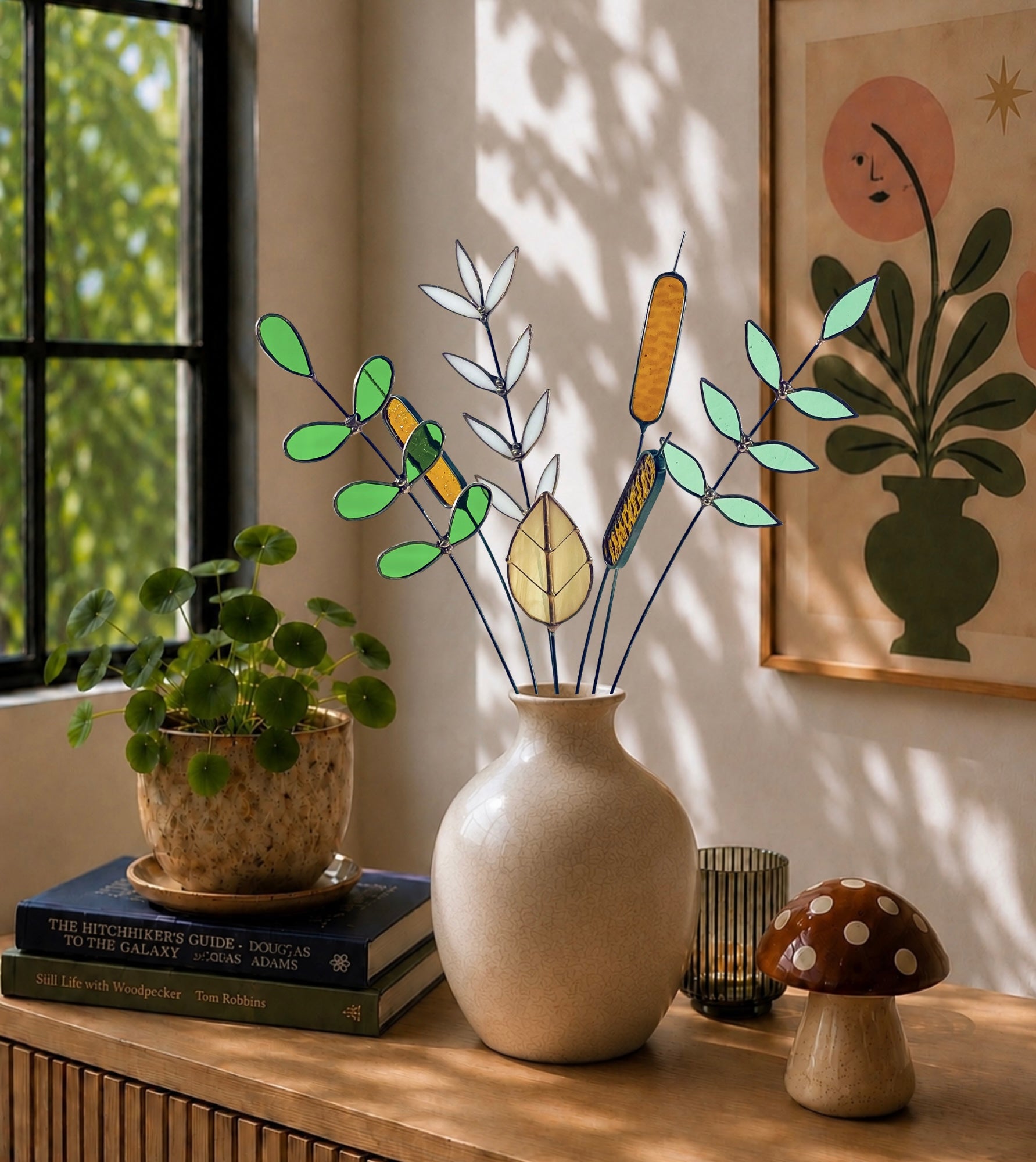 A stained glass plant arrangement with green, yellow, and white leaves and cattails in a beige ceramic vase, styled on a wooden table with a potted plant, books, a candle holder, and a small mushroom decor piece in a sunlit room.