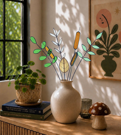 A stained glass plant arrangement with green, yellow, and white leaves and cattails in a beige ceramic vase, styled on a wooden table with a potted plant, books, a candle holder, and a small mushroom decor piece in a sunlit room.