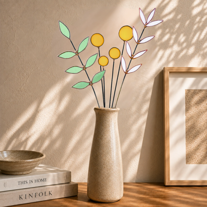 A ceramic vase with a textured beige finish holding a decorative stained glass botanical arrangement of yellow billy balls and pale green leaves on thin black stems, styled on a wooden surface beside stacked books and a framed print against a warm beige wall with soft shadow patterns.
