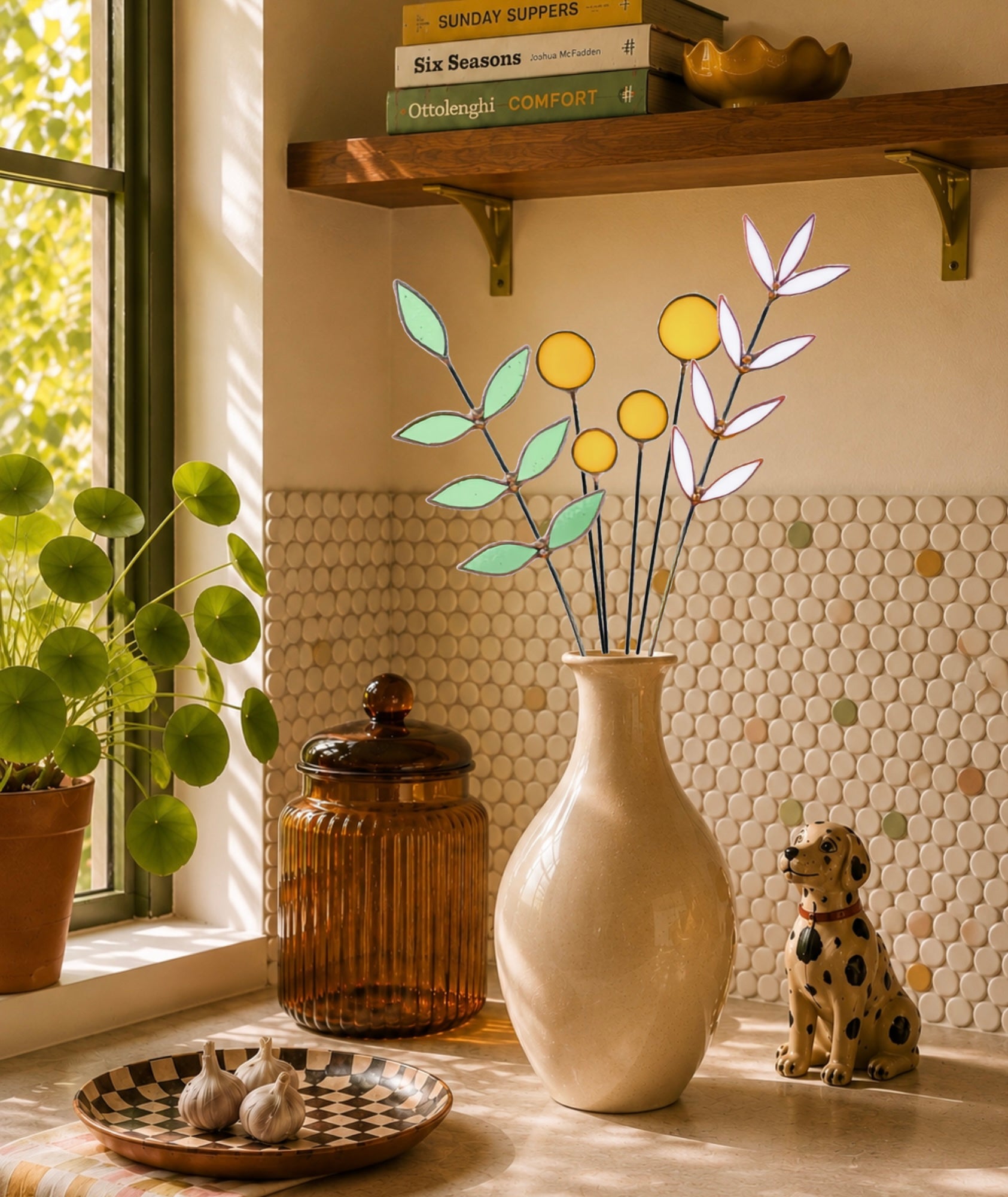 A decorative stained glass botanical arrangement with yellow circles and green leaves displayed in a ceramic vase on a kitchen counter next to a dog statue and a window.