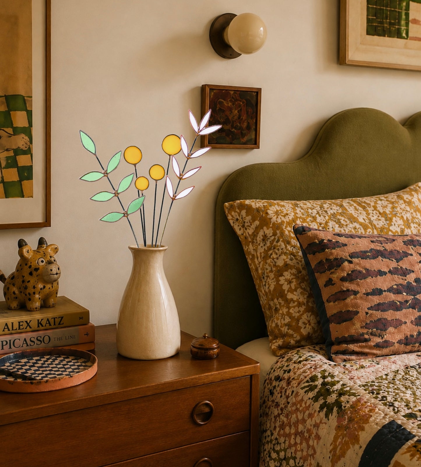 A decorative stained glass botanical arrangement displayed in a beige ceramic vase on a wooden bedside table in a styled bedroom.