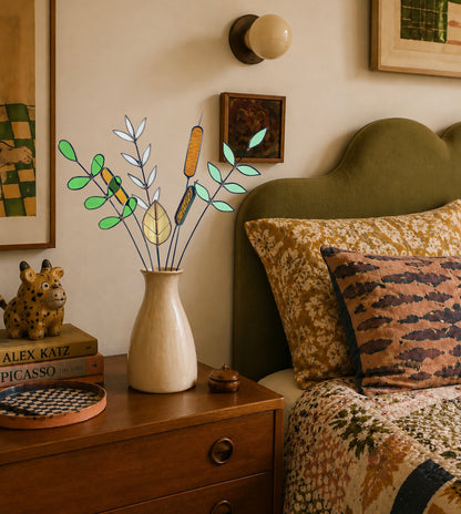 A styled bedroom scene with a beige ceramic vase holding a decorative dried plant arrangement with green leaves and tan accents, placed on a wooden nightstand beside a bed with patterned pillows.