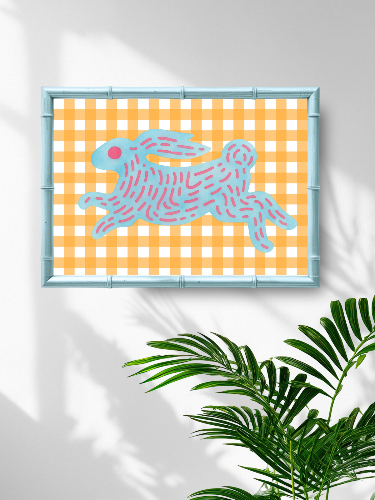 Framed artwork with a blue rabbit on an orange checkered background, placed on a white wall with a plant in the foreground.
