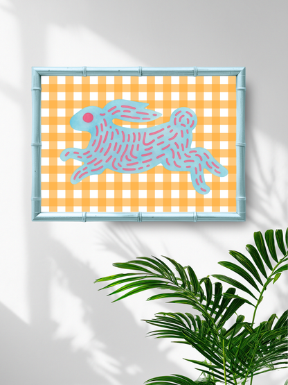 Framed artwork with a blue rabbit on an orange checkered background, placed on a white wall with a plant in the foreground.