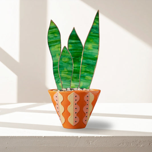 Handmade stained glass snake plant with green glass leaves arranged in a patterned orange planter, styled on a sunlit shelf.