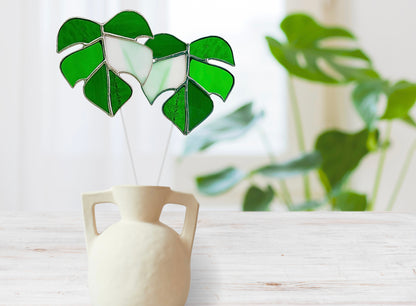 Decorative green leaf sculpture in a white vase on a light wooden surface with a blurred plant in the background.
